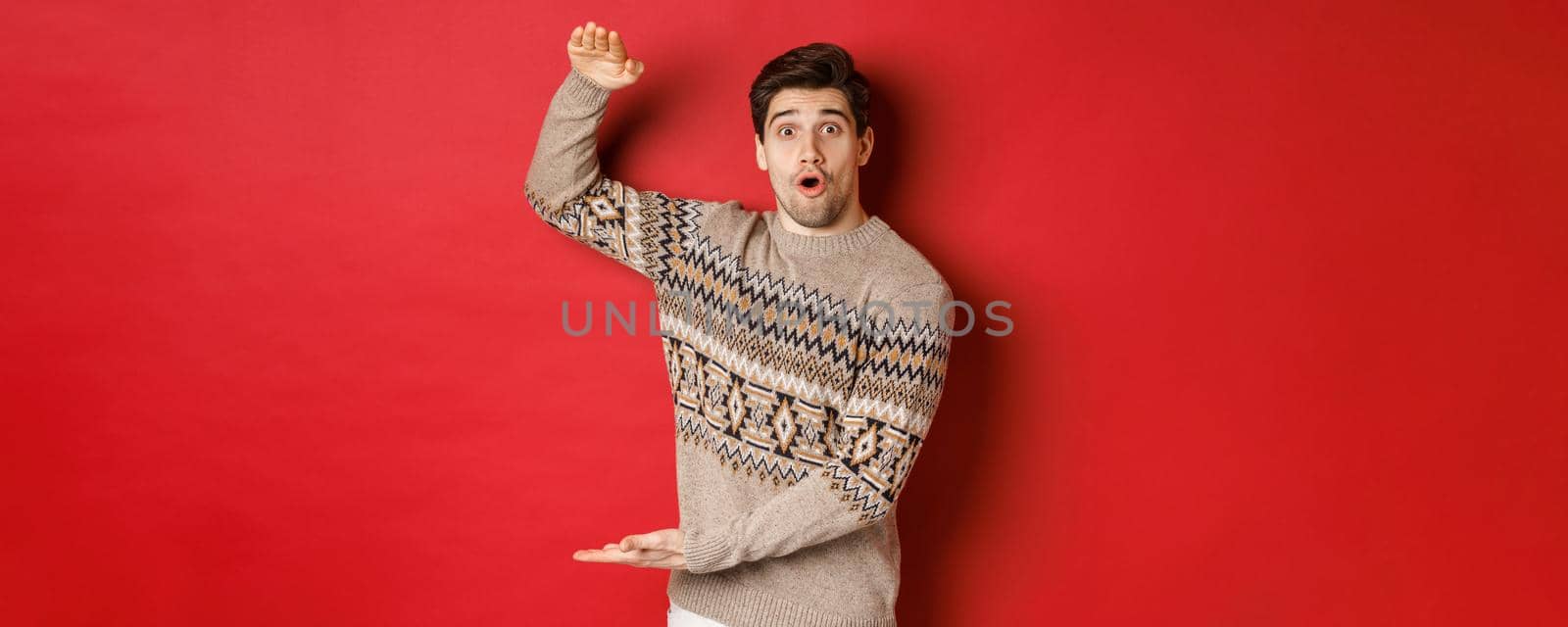 Image of attractive man in christmas sweater, shaping big new year gift, showing something large and amazing, standing over red background by Benzoix