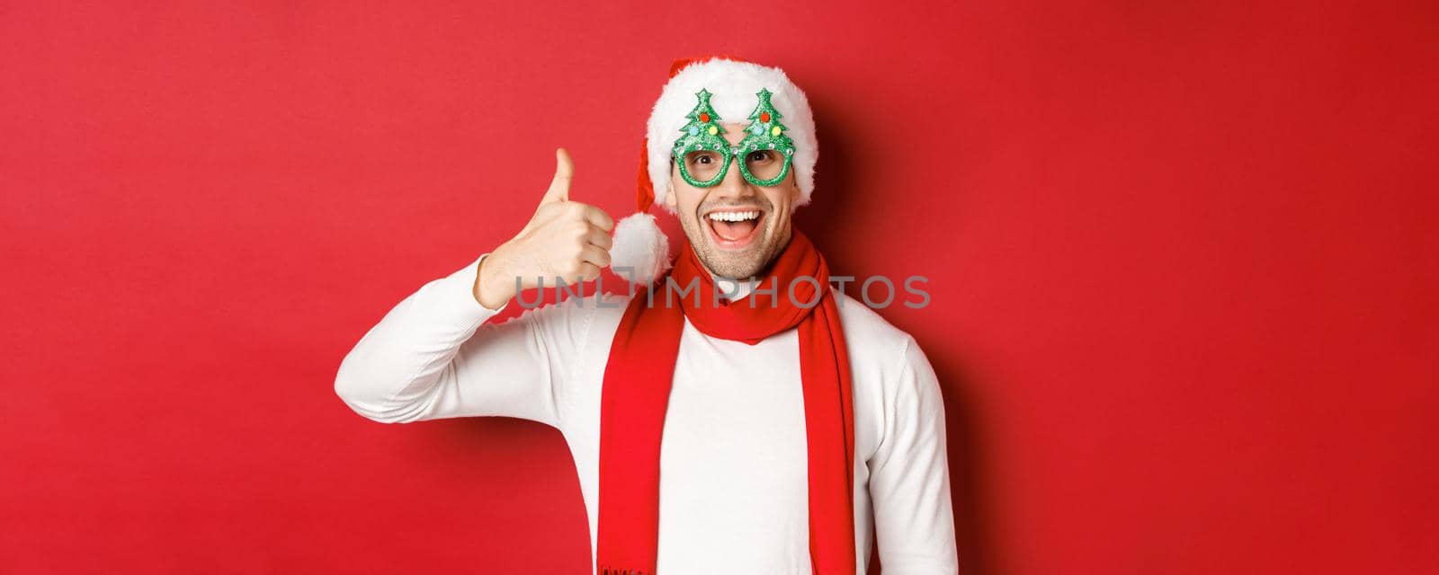 Concept of christmas, winter holidays and celebration. Cheerful guy in santa hat and party glasses, showing thumb-up and smiling pleased, standing over red background by Benzoix