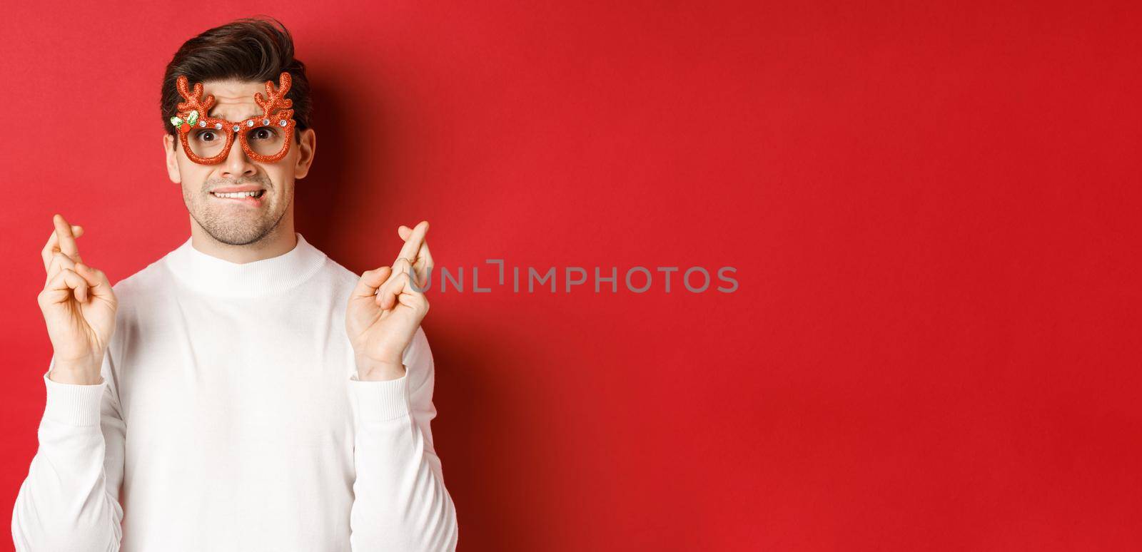 Close-up of handsome nervous guy in party glasses, making a wish, crossing fingers for good luck and looking with hope at camera, standing over red background by Benzoix