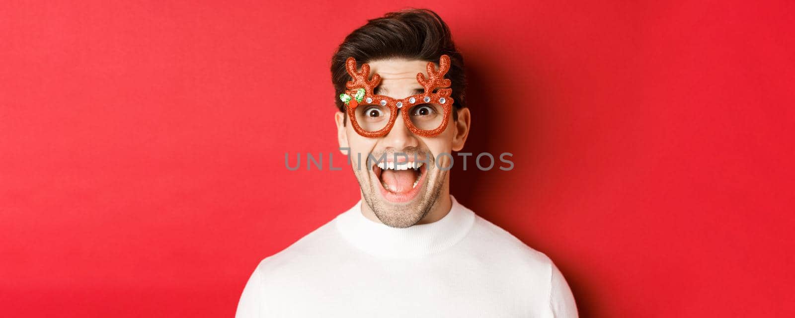 Concept of winter holidays, christmas and celebration. Close-up of attractive guy with bristle, wearing new year party glasses and looking amazed at promo offer, standing over red background by Benzoix