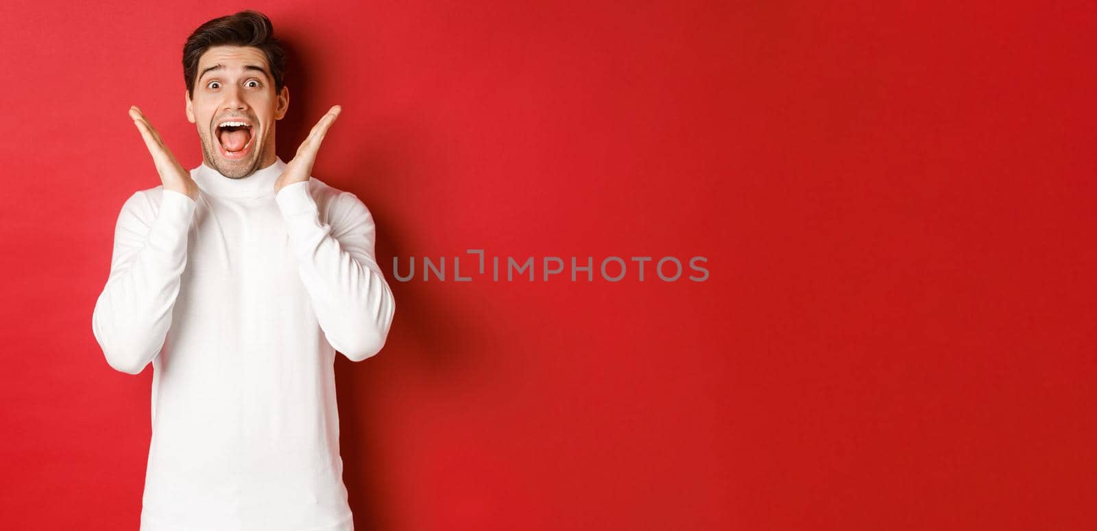 Concept of winter holidays, christmas and lifestyle. Surprised handsome man in white sweater, reacting to amazing new year promo offer, screaming amazed and raising hands, red background by Benzoix