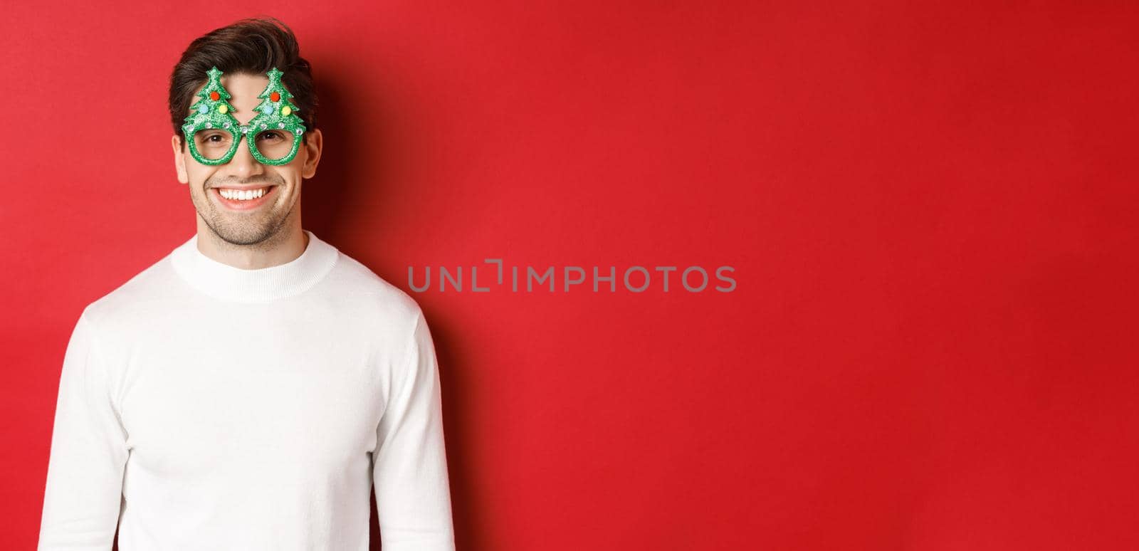 Concept of christmas, winter holidays and celebration. Close-up of handsome man in white sweater and party glasses, enjoying new year, standing over red background by Benzoix