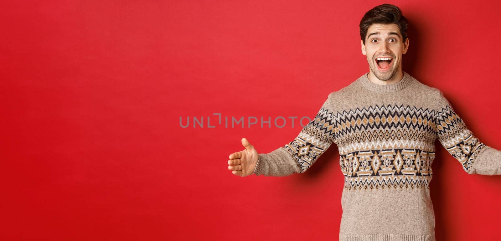 Portrait of attractive happy man in christmas sweater showing big present, spread hands sideways as if holding large box, standing over red background by Benzoix