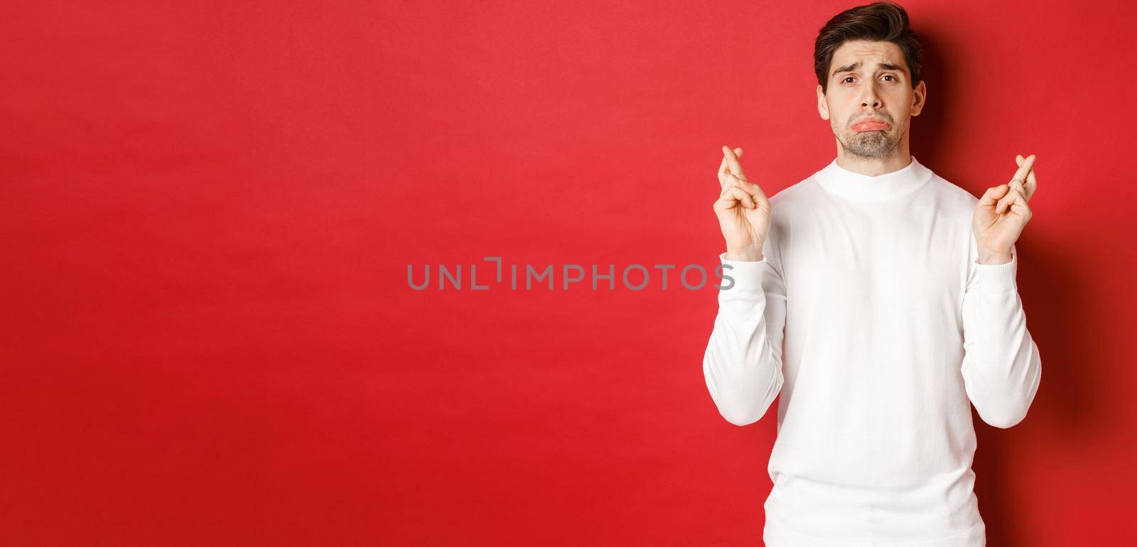 Image of sad and gloomy guy in white sweater, crying with fingers crossed, waiting for something or praying, standing over red background.
