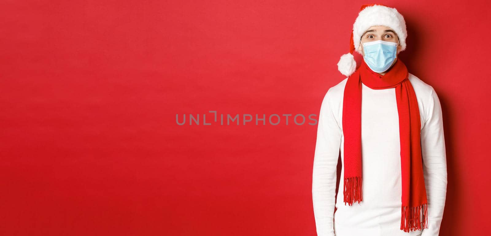 Concept of new year, coronavirus and holidays. Cheerful man celebrating christmas and social distancing, wearing medical mask, santa hat and scarf, standing over red background.