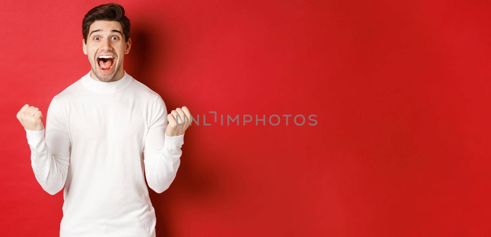 Image of happy good-looking man in white sweater, winning something, making fist pump and smiling amazed, celebrating victory, standing over red background.