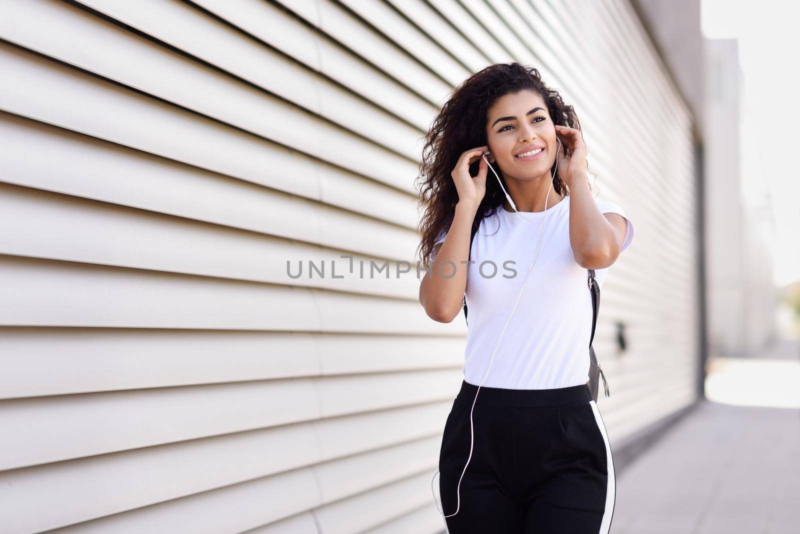 Funny African woman listening to music and dancing with earphones and smartphone outdoors. Arab girl in sport clothes with curly hairstyle in urban background.