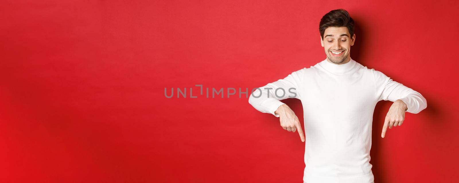 Image of dreamy good-looking man in white sweater, looking and pointing fingers down at copy space, advertisement about new year and holidays, standing over red background.