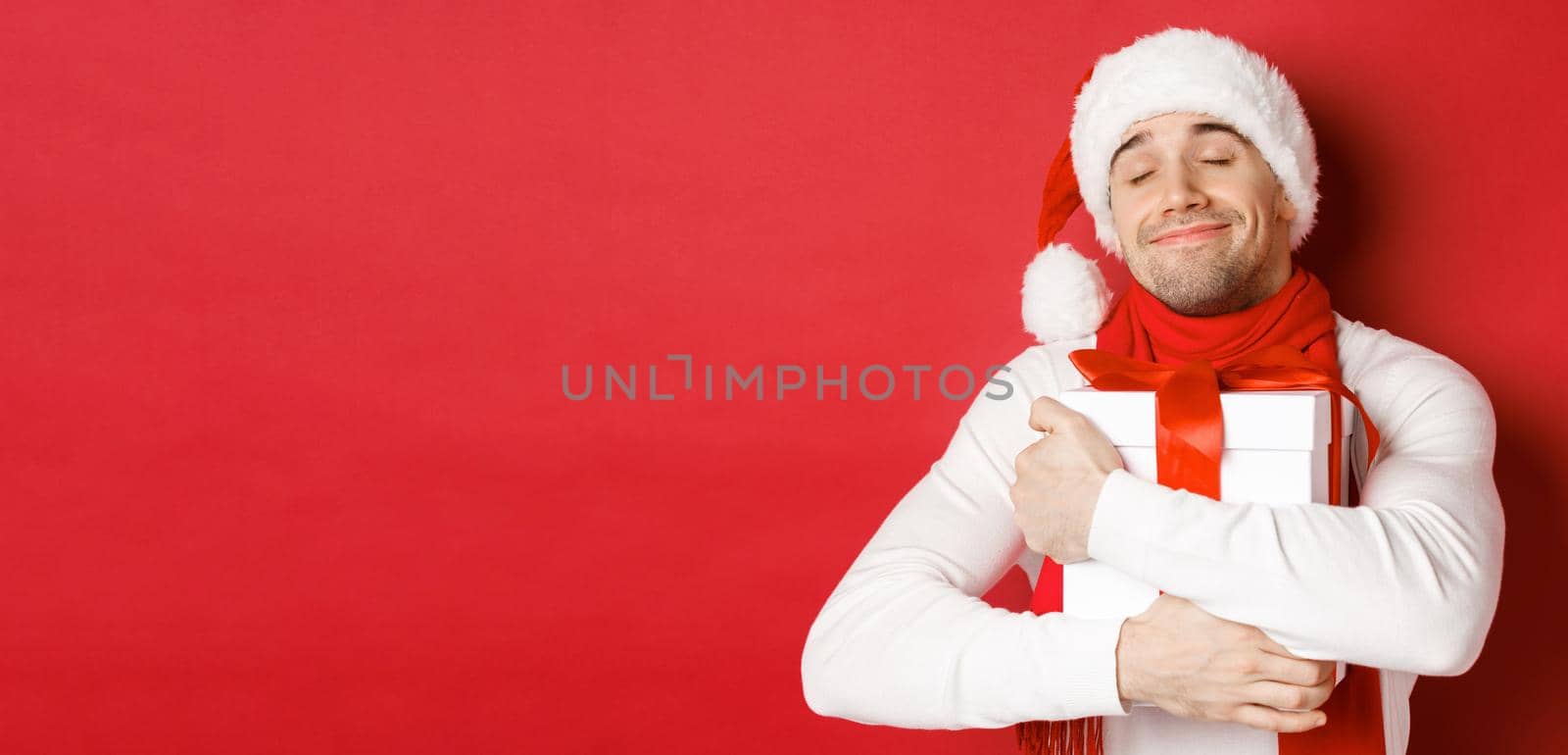 Concept of winter holidays, christmas and lifestyle. Image of lovely guy in santa hat and scarf, hugging his new year present and smiling flattered, standing over red background. Copy space