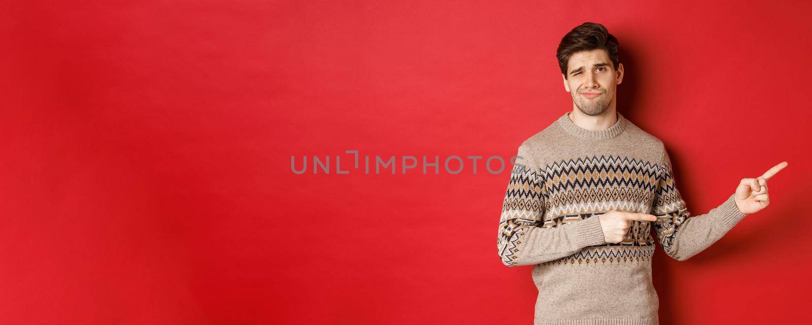 Concept of christmas celebration, winter holidays and lifestyle. Image of skeptical and displeased handsome man in xmas sweater, pointing fingers right and do not recommend product.