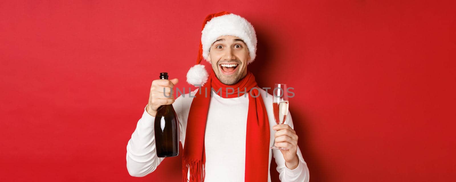 Concept of winter holidays, christmas and lifestyle. Close-up of cheerful handsome man, holding champagne bottle and glass, celebrating new year, standing over red background by Benzoix