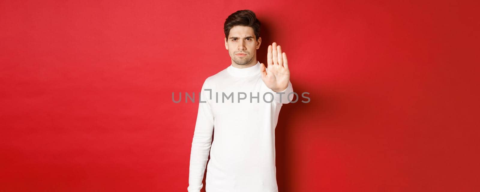 Image of serious and confident man telling to stop, forbid something, extending one hand and prohibit action, standing over red background.