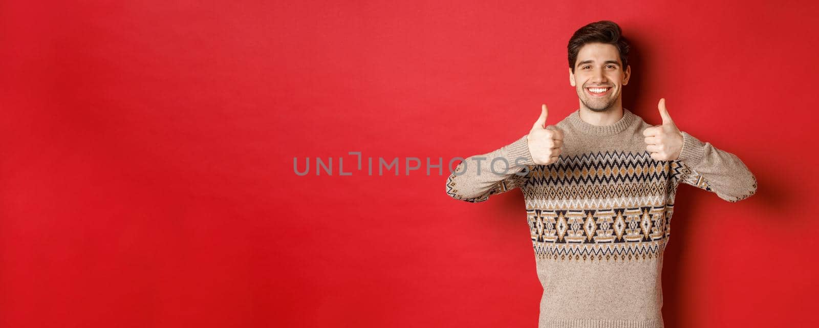 Portrait of happy and pleased handsome guy in christmas sweater, showing thumbs-up and nod in approval, smiling satisfied, standing over red background by Benzoix