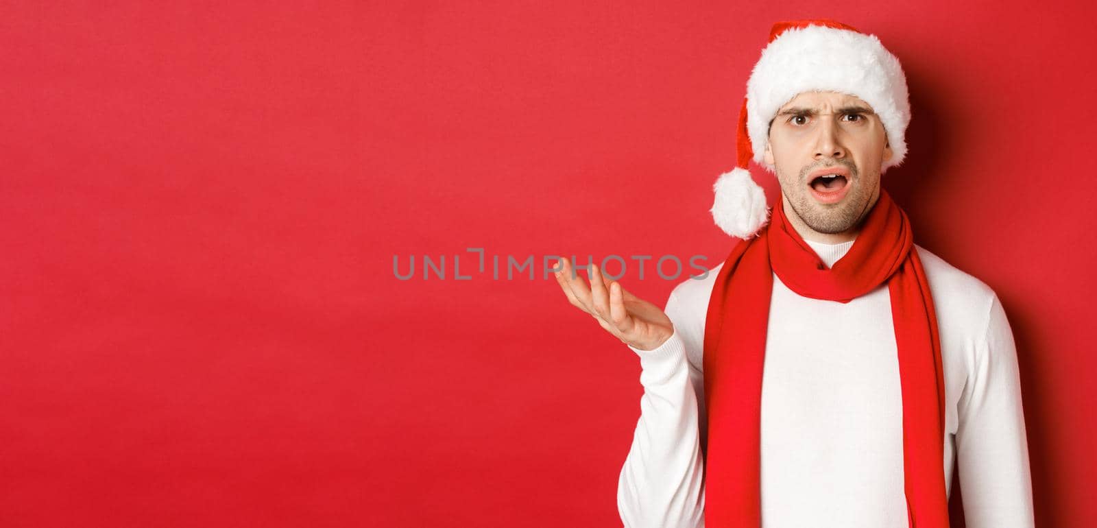 Concept of christmas, winter holidays and celebration. Portrait of confused man in santa hat and scarf, frowning and looking perplexed, standing over red background.