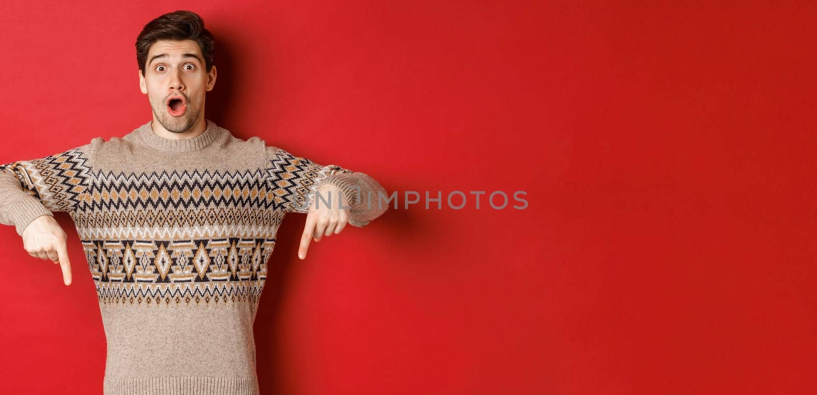 Concept of christmas celebration, winter holidays and lifestyle. Impressed handsome man in xmas sweater, pointing fingers down with amazed face, standing over red background by Benzoix