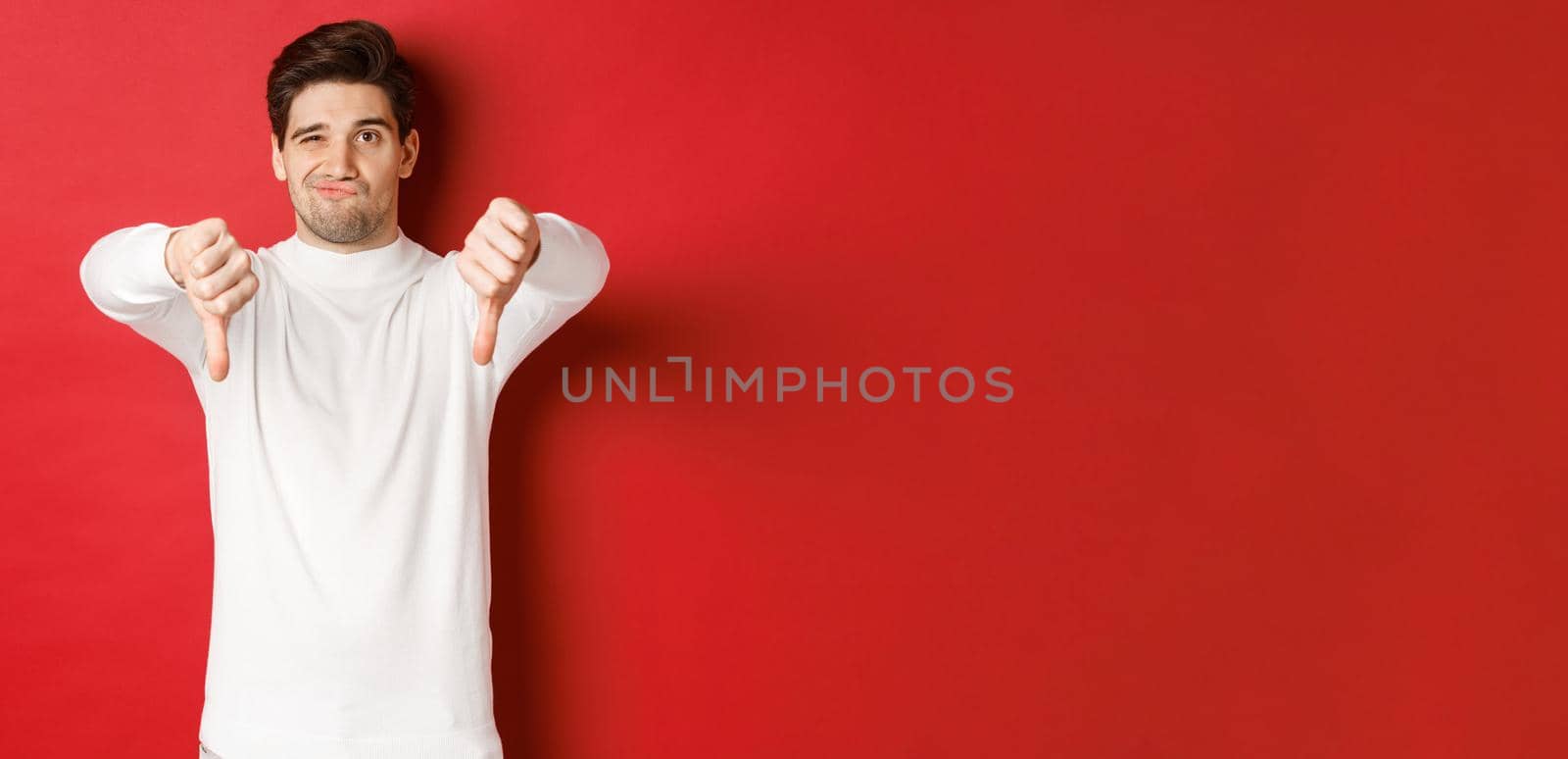 Portrait of disappointed skeptical guy, wearing white sweater, showing thumbs-down to disagree, express dislike, standing over red background by Benzoix