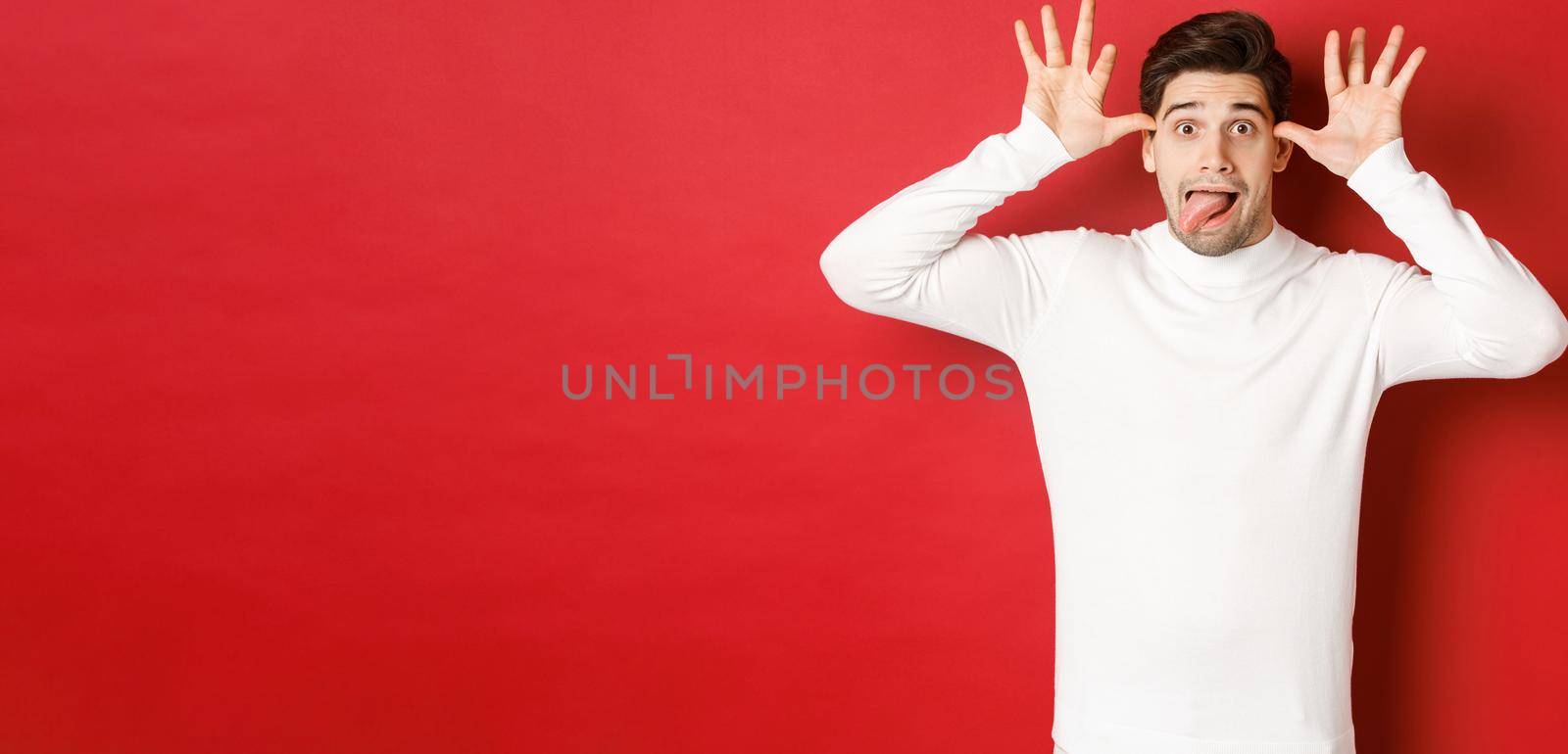 Portrait of funny caucasian guy, showing tongue and making faces, wearing white sweater, standing against red background.