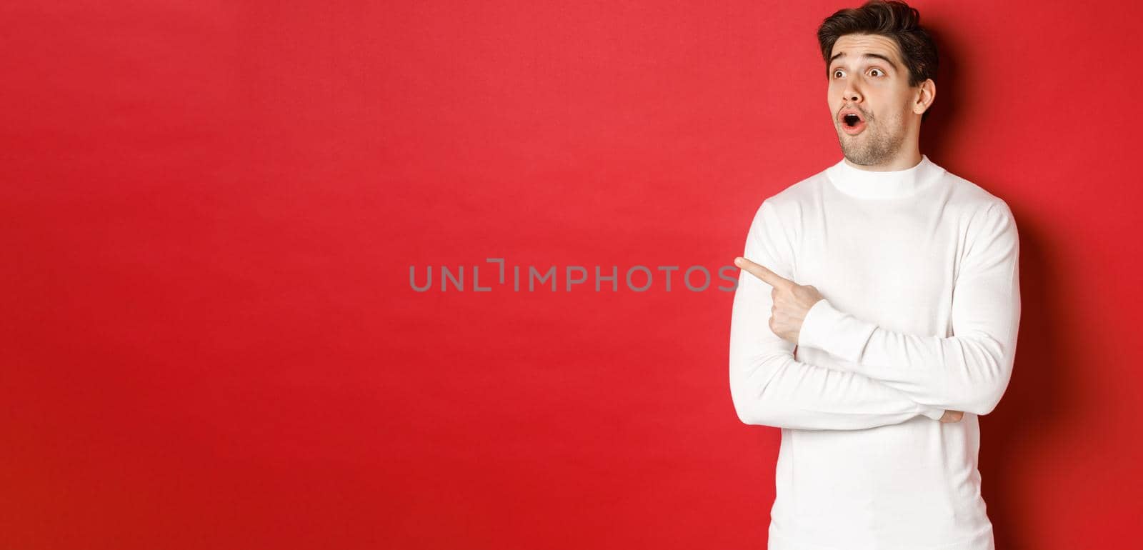 Concept of winter holidays. Portrait of impressed handsome man in white sweater, pointing and looking left with dropped jaw and amazed expression, standing against red background by Benzoix