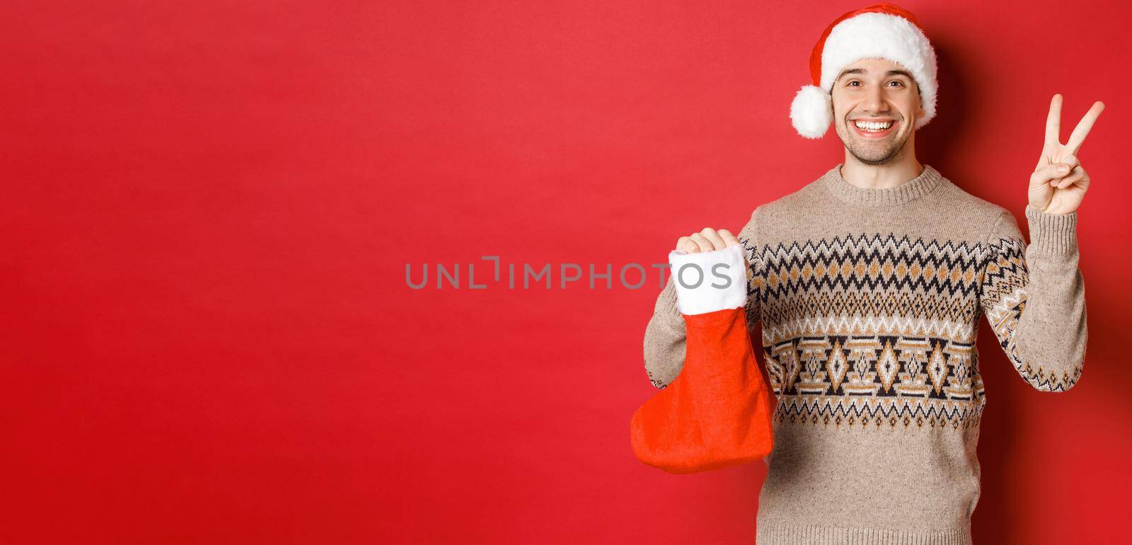 Concept of winter holidays, new year and celebration. Image of happy smiling man in santa hat and sweater, showing peace sign and a christmas stocking bag with gifts, red background.