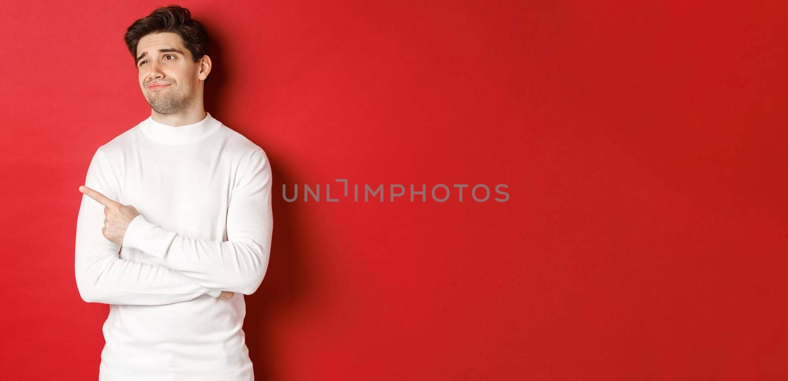 Concept of winter holidays. Image of displeased young man in white sweater, looking and pointing finger left at something average, standing skeptical over red background by Benzoix