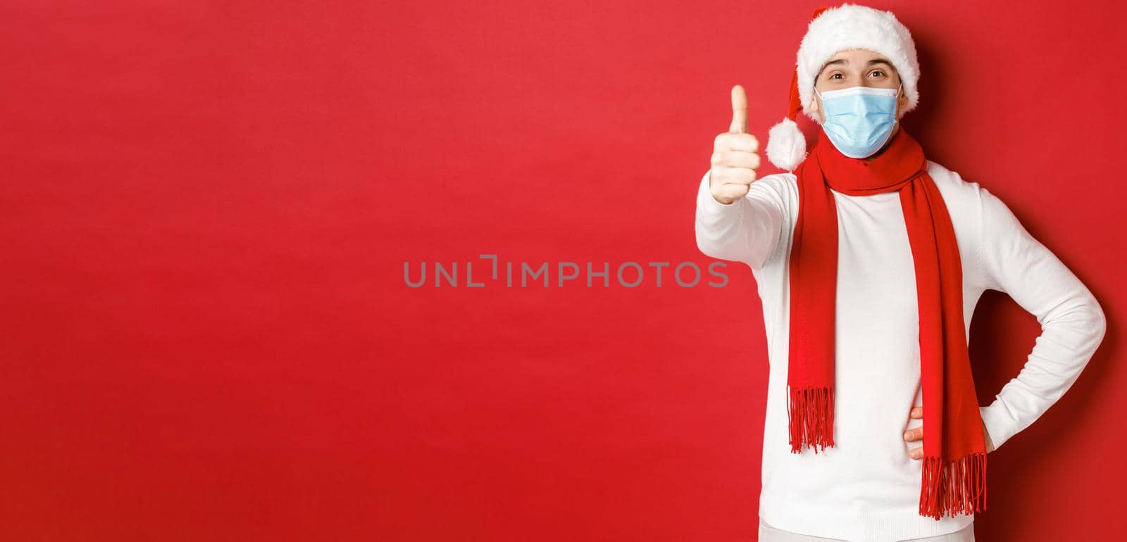 Concept of covid-19, christmas and holidays during pandemic. Cheerful handsome man in medical mask and santa hat, showing thumb-up, celebrating new year and social distancing.