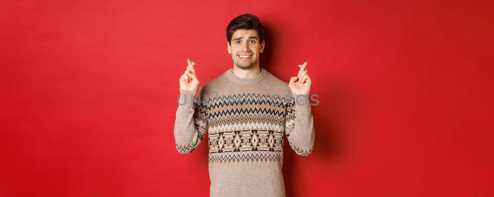 Image of hopeful and worried man in christmas sweater waiting for something, cross fingers for good luck and making wish, nervous about new year gift, red background by Benzoix