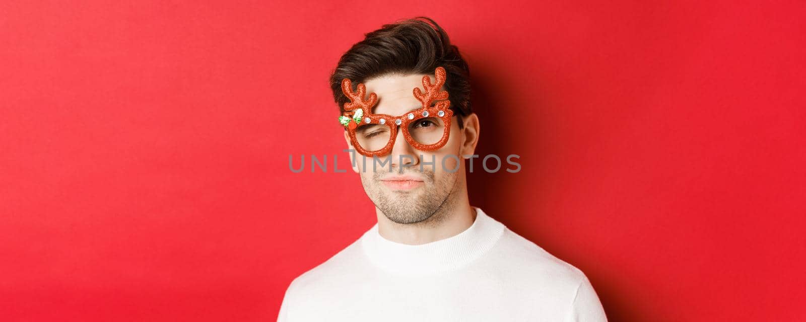 Concept of winter holidays, christmas and celebration. Close-up of cheeky attractive man in party glasses, winking and looking sassy, standing over red background by Benzoix