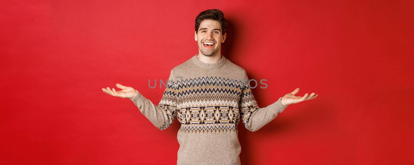 Portrait of happy good-looking man celebrating new year holidays, wearing christmas sweater, spread hands sideways and smiling, holding something on copy space, standing over red background.