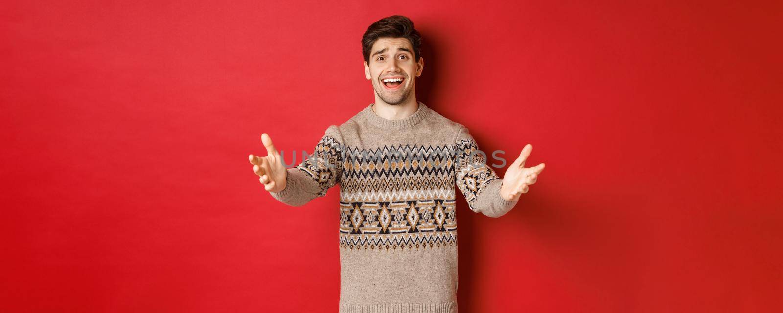 Image of happy and flattered handsome guy in christmas sweater, reaching hands to welcome guests on new year party, inviting to celebration, standing over red background by Benzoix