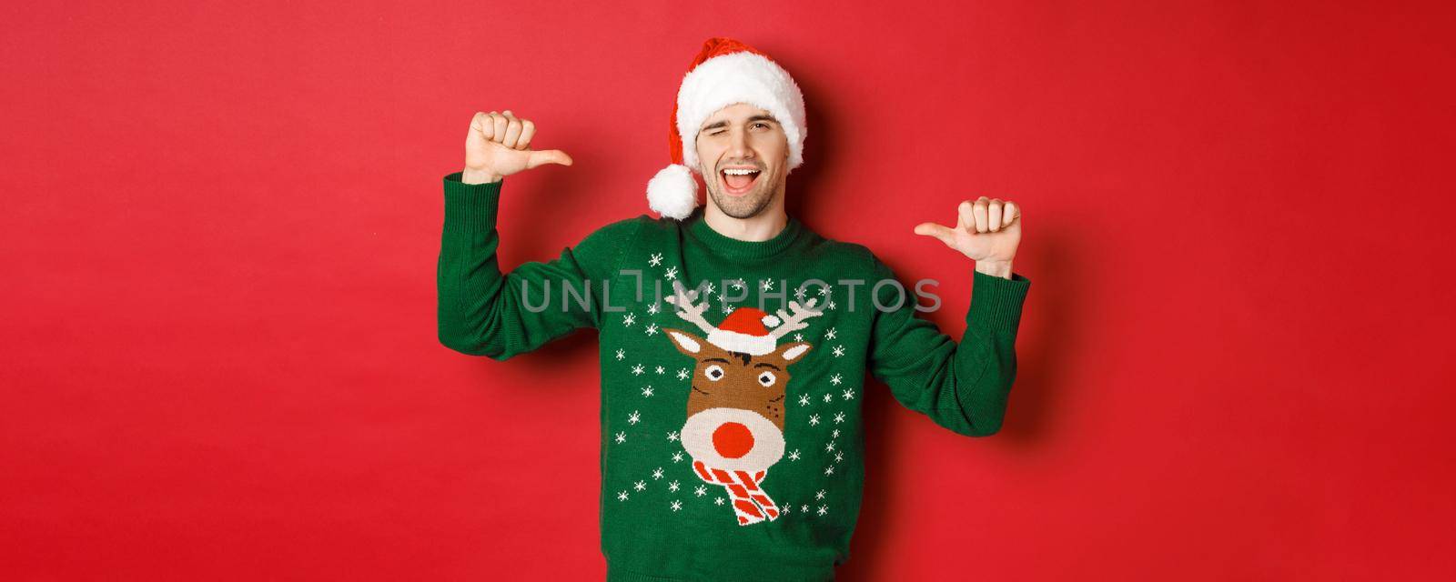 Concept of winter holidays, christmas and lifestyle. Sassy handsome man in santa hat and green sweater, pointing at himself and winking, standing over red background.