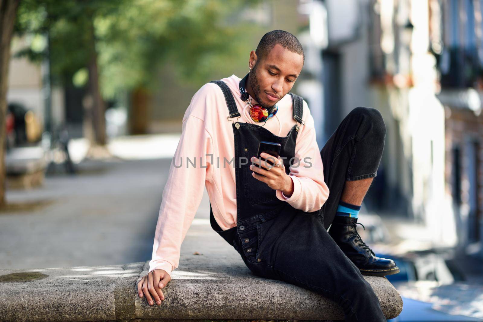 Young black man with headphones sitting in urban street looking at his smart phone. Lifestyle concept.