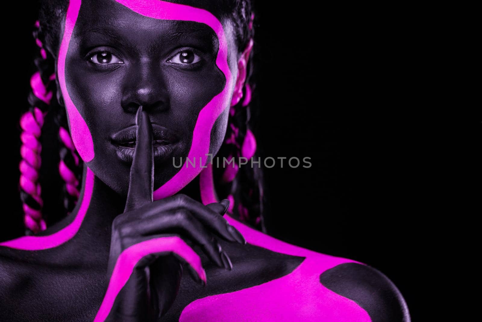 Face art. Girl showing gesture quietly, asks remain silent with finger. Woman with black and pink body paint. Young african girl with colorful bodypaint. An amazing afro american model with makeup