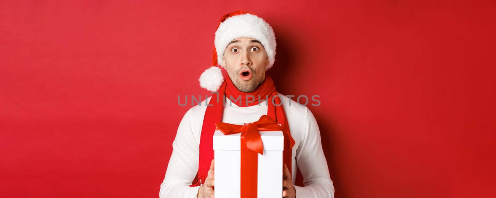 Concept of winter holidays, christmas and lifestyle. Close-up of surprised handsome guy in santa hat and scarf, looking amazed and holding new year gift, standing over red background by Benzoix