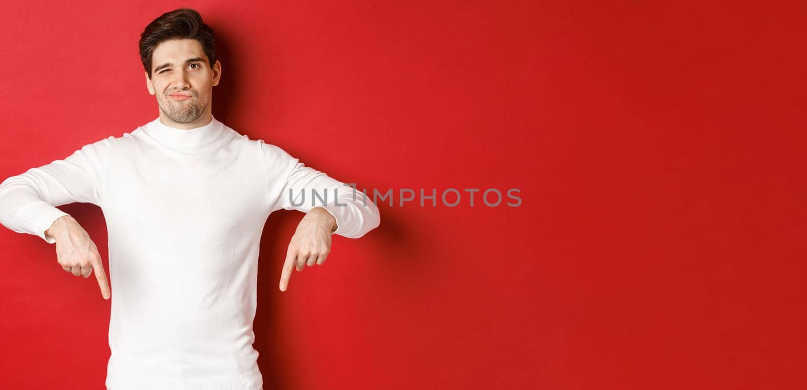 Portrait of attractive, skeptical young man in white sweater, grimacing and pointing fingers down, disagree and dislike something, standing over red background by Benzoix