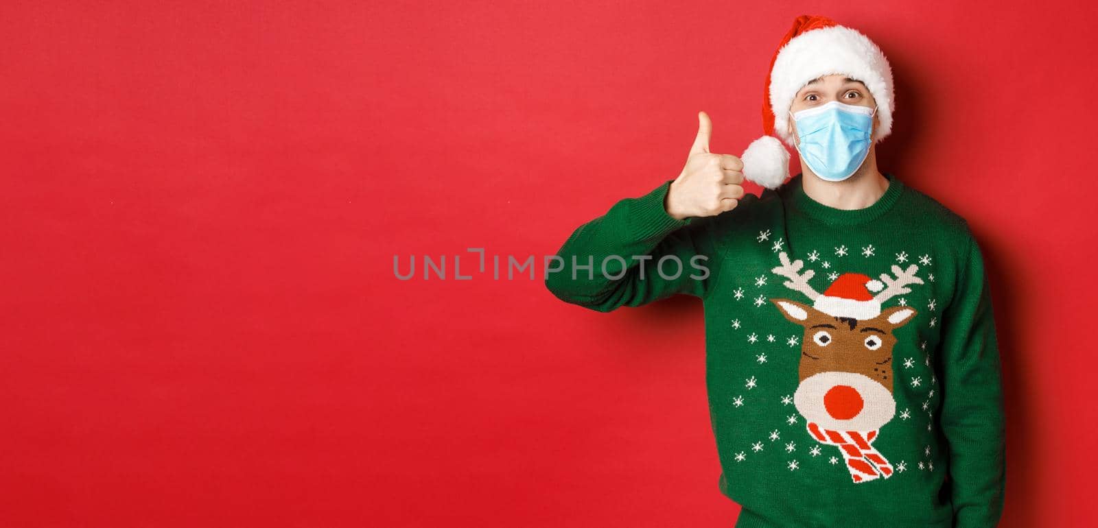 Concept of new year, covid-19 and social distancing. Amazed young guy celebrating christmas, wearing medical mask and santa hat, showing thumb-up in approval, red background by Benzoix