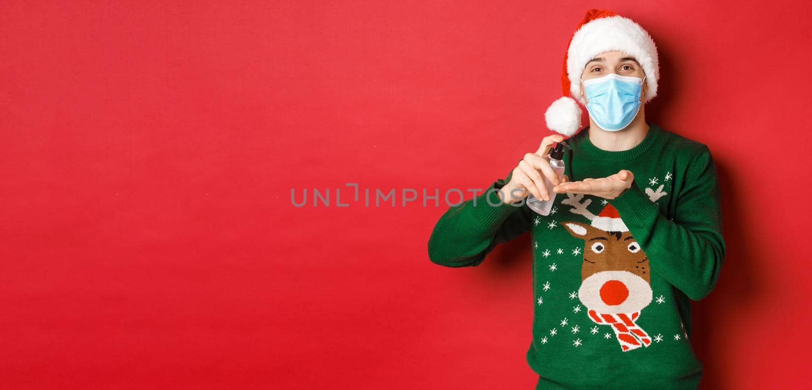 Concept of new year, coronavirus and social distancing. Attractive young man in santa hat, medical mask and christmas sweater, using hand sanitizer to clean hands, standing over red background.
