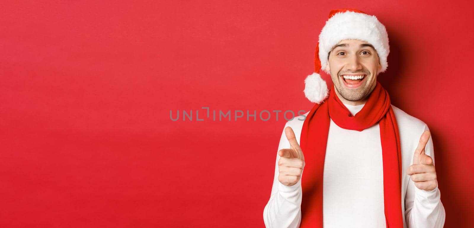 Concept of christmas, winter holidays and celebration. Cheeky man in santa hat and scarf, smiling and pointing fingers at camera, wishing happy new year, standing over red background.