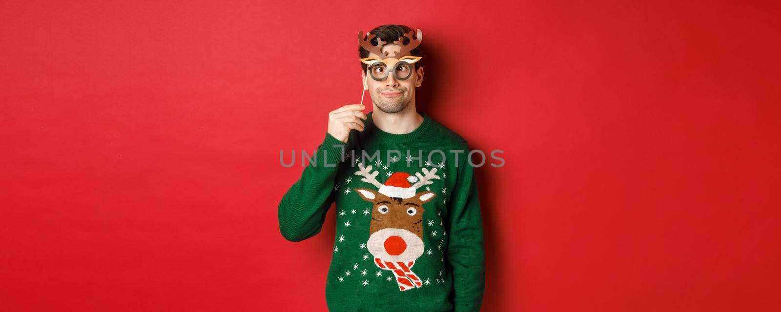 Funny man in christmas sweater and party mask, celebrating winter holidays, showing funny faces, standing against red background by Benzoix