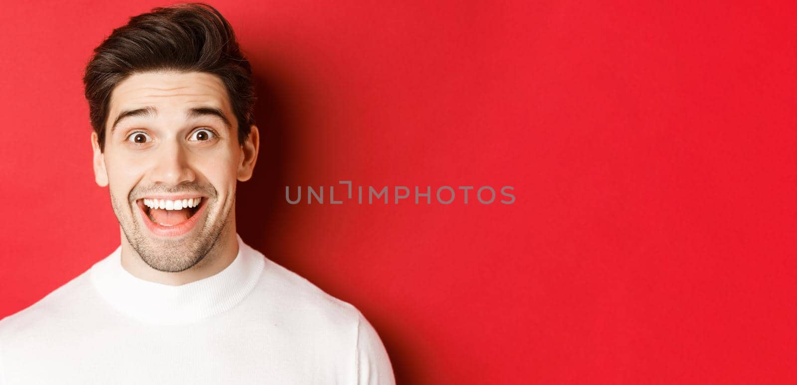 Close-up of surprised happy guy in white sweater, looking amused, hear interesting news, standing over red background.