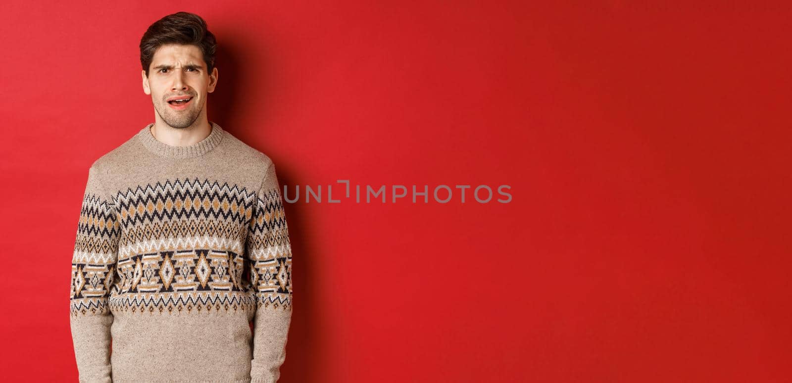 Portrait of confused adult man in christmas sweater, standing disappointed during new year holidays, dont understand something, standing over red background.