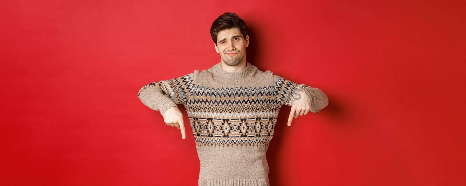 Concept of christmas celebration, winter holidays and lifestyle. Skeptical and unamused handsome man, showing something bad, pointing fingers down and grimacing, red background.