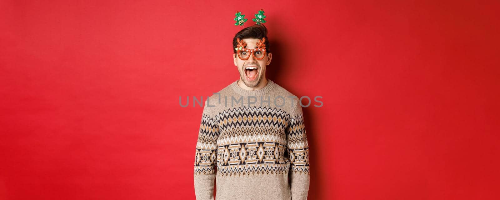 Image of surprised and excited handsome man, wearing party glasses with christmas sweater, drop jaw and looking at amazing new year promo offer, standing against red background.