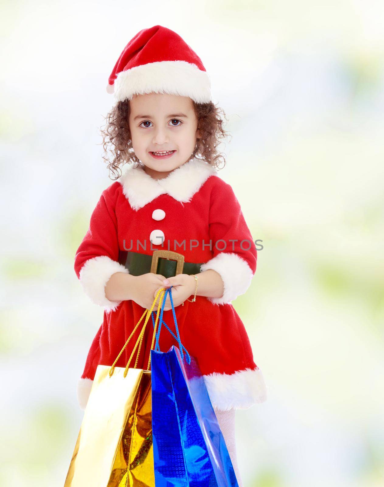 Gentle little curly-haired girl in a coat and hat of Santa Claus holding colorful shopping bags. Close-up.white-green blurred abstract background with snowflakes.