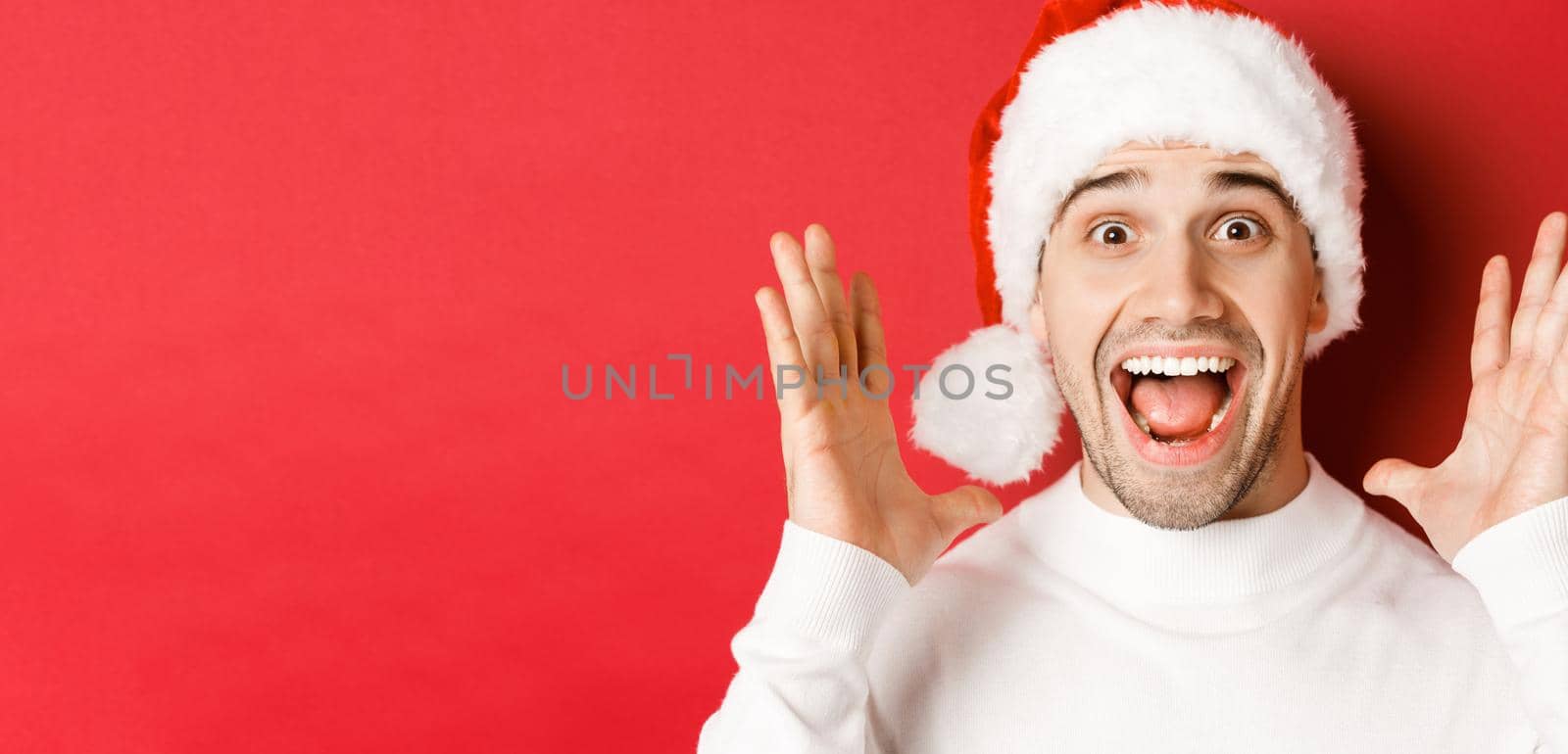 Close-up of happy young man in santa hat, making big christmas announcement, smiling amazed, standing over red background.