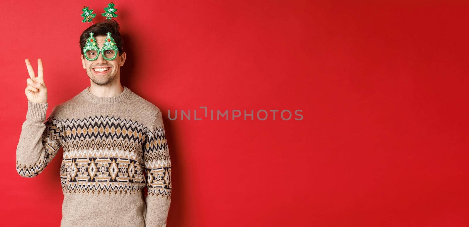 Portrait of funny and cute man in party glasses and christmas sweater, smiling and showing peace sign while posing over red background for new year advertisement.