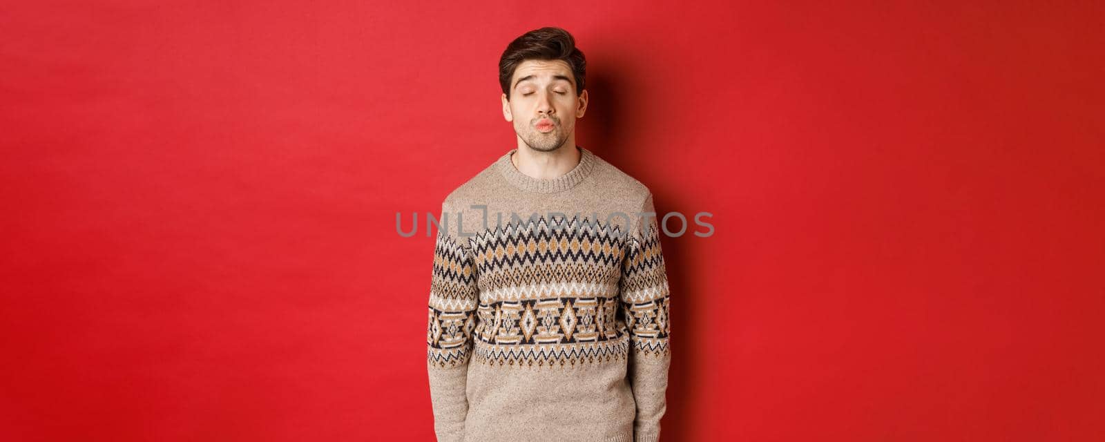 Image of handsome man in christmas sweater pucker lips and close eyes, waiting for kiss under mistletoe, standing over red background.