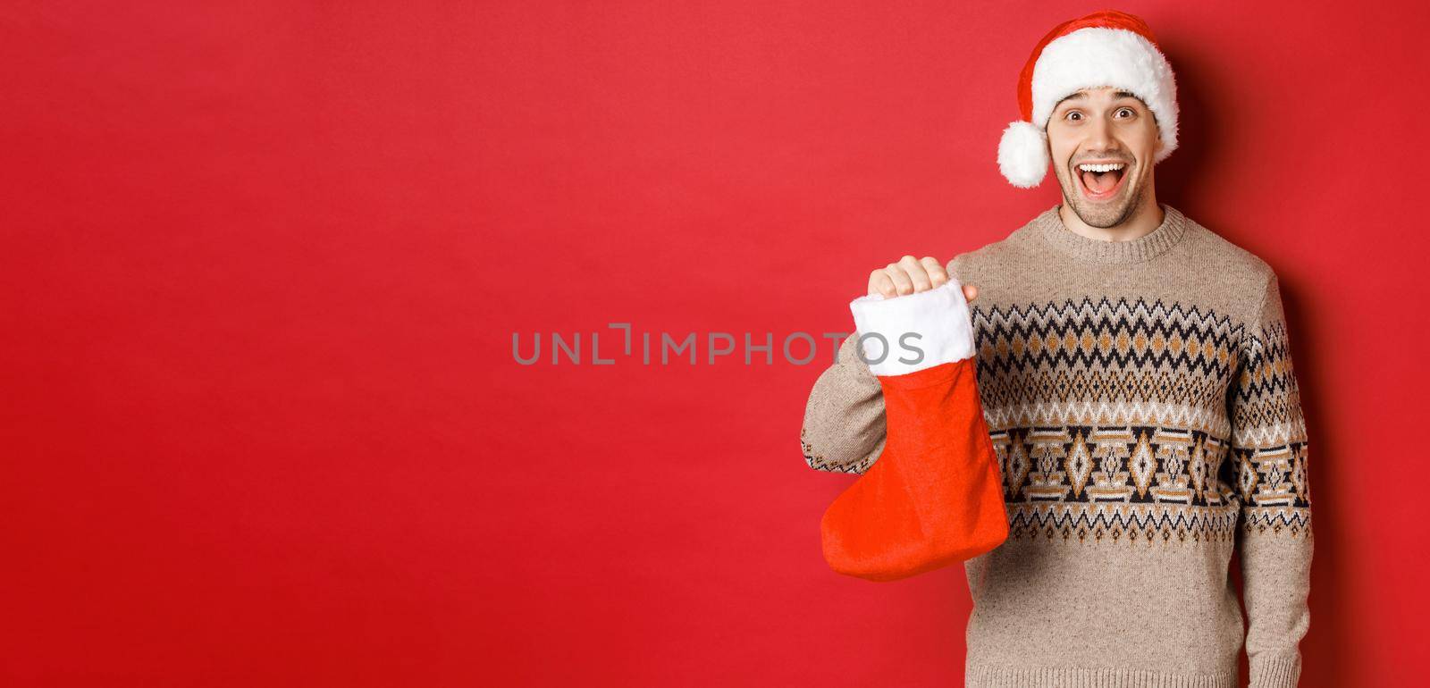 Concept of winter holidays, new year and celebration. Cheerful and surprised adult man receiving candies on Saint Nicholas day in red stocking, standing amazed over red background by Benzoix