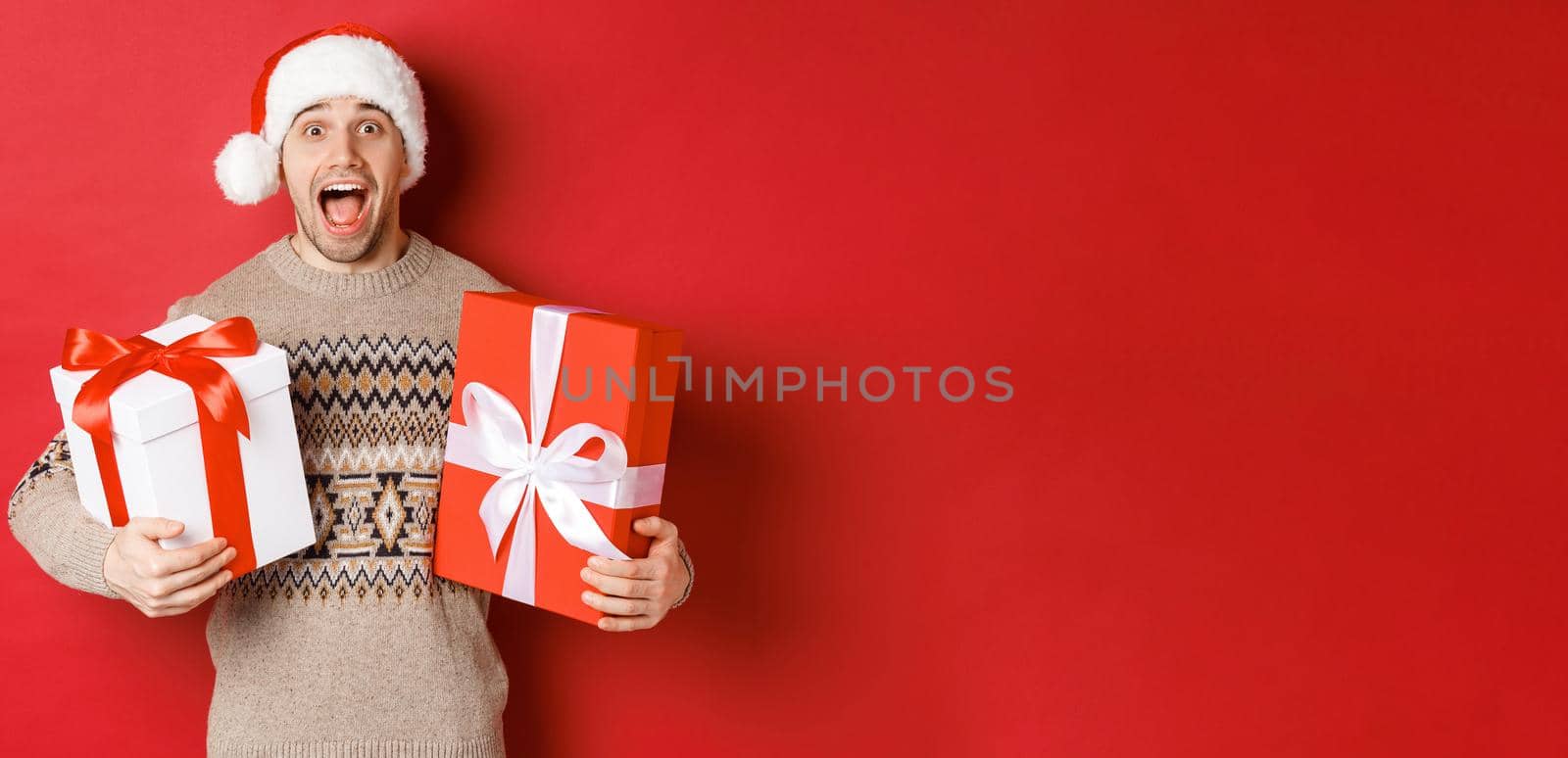 Image of cheerful attractive guy holding christmas presents, standing in santa hat and winter sweater, smiling amazed, standing over red background.