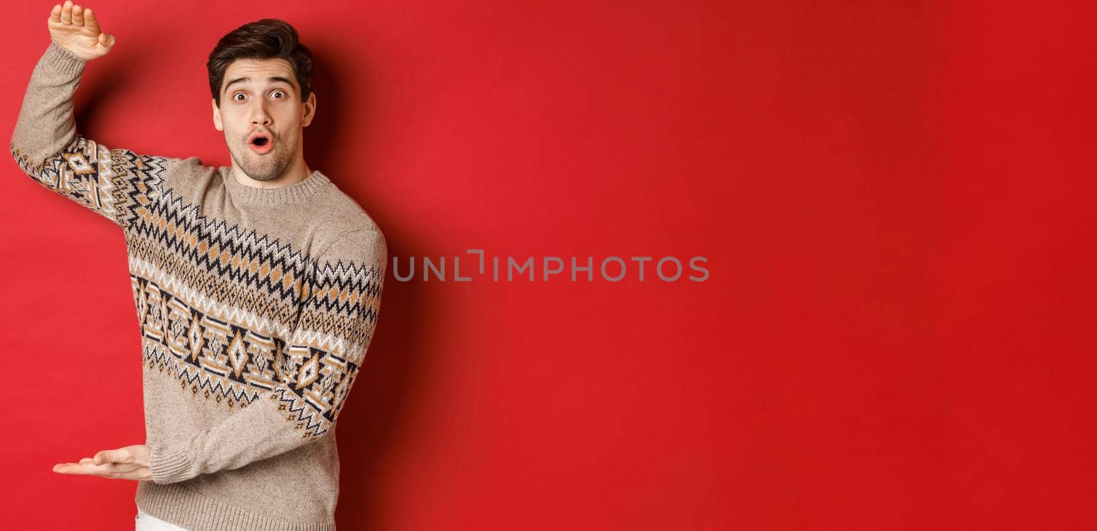 Image of attractive man in christmas sweater, shaping big new year gift, showing something large and amazing, standing over red background.