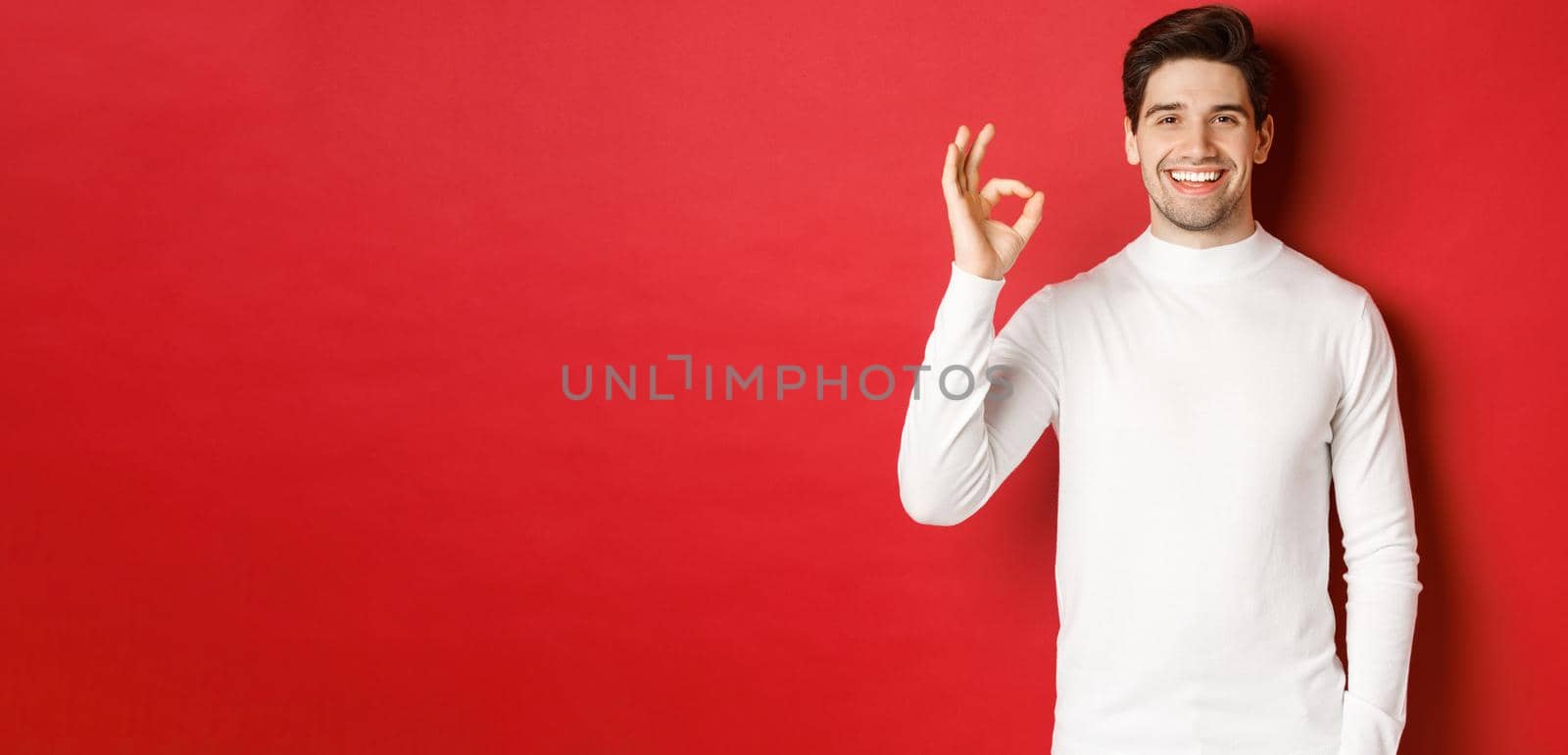 Concept of winter holidays, christmas and lifestyle. Happy handsome man in white sweater showing okay sign, smiling pleased, recommending shop, standing over red background.